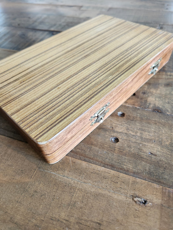 A closed, folding wooden backgammon set, featuring small metal latch closures, resting on a rustic wooden surface.
