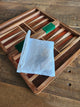 A wooden backgammon board with striped playing surfaces and two green dice, shown open on a rustic wooden table. A white mesh drawstring pouch is placed in front of the board showing the bag that the playing pieces come in.