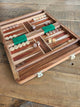 An open wooden backgammon board set with inlaid wooden playing surface, green felt, doubles block, checkers, and dice, placed on a rustic wooden tabletop.