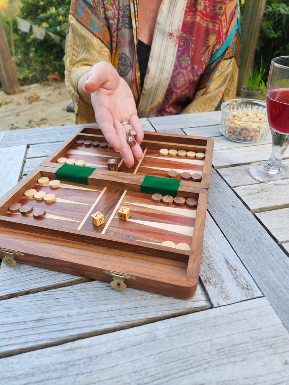 A wooden backgammon board with wooden pieces set on a weathered gray wooden table outdoors. A person's hand is visible holding a pair of dice, with a glass of red wine and a bowl of nuts on the table nearby.