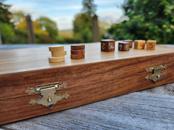 A wooden board game set with stacked round tokens and numbered cube blocks placed on a polished wooden box, photographed outdoors with a blurred natural background.
