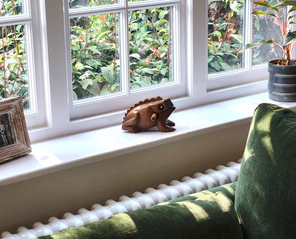 Wooden frog figurine on a windowsill with greenery outside