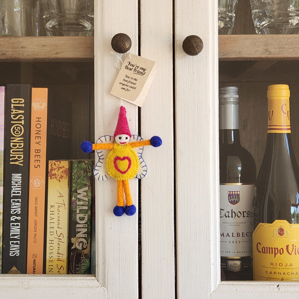 A small handmade felt 'best friend' keepsake ornament with a pink cone hat, orange body, blue pom-pom hands and feet, and a yellow heart on the front, hanging on a white cabinet door.