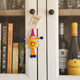 A small handmade felt 'best friend' keepsake ornament with a pink cone hat, orange body, blue pom-pom hands and feet, and a yellow heart on the front, hanging on a white cabinet door.