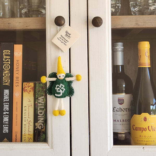 A small handmade felt fairy/pixie keepsake with a yellow cone hat, green dress, and yellow shoes hangs on a white cabinet door. A gift tag with a 'Good Luck' message is attached.