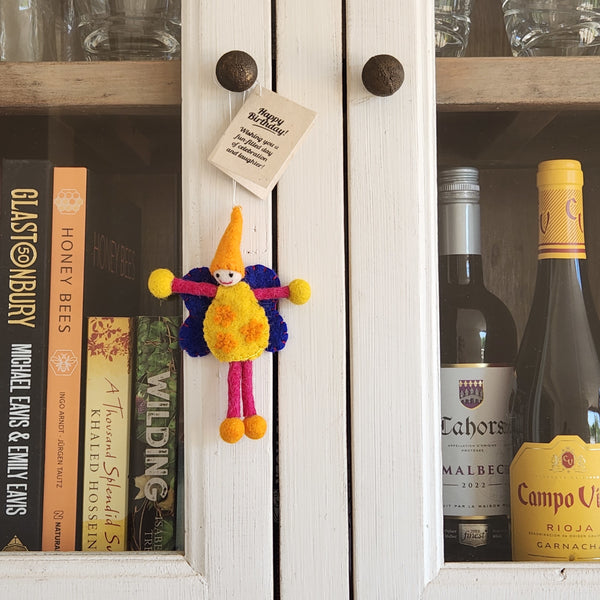 A small handmade felt fairy/pixie keepsake with a yellow body, orange hat, blue scarf, and pink legs hangs on a white wooden cabinet door with a 'Happy Birthday!' note.