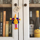 A small handmade felt fairy/pixie keepsake with a yellow body, orange hat, blue scarf, and pink legs hangs on a white wooden cabinet door with a 'Happy Birthday!' note.
