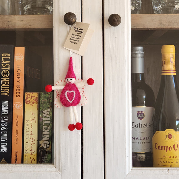 A small pink felt fairy/pixie keepsake with a white heart on its belly, hanging on a white wooden cabinet door.