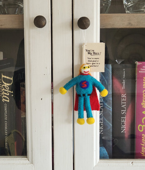 A small handmade felt superhero keepsake/gift tag with a 'You're My Hero!' tag hanging on a white cabinet door in front of books.