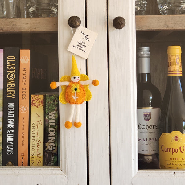 A small handmade felt fairy/pixie keepsake with orange and yellow colours hangs on a white wooden cabinet door next to a bookshelf.