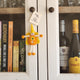 A small handmade felt fairy/pixie keepsake with orange and yellow colours hangs on a white wooden cabinet door next to a bookshelf.