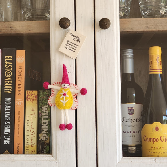 A small handmade felt fairy/pixie with a pink hat, yellow dress, and pink pom-pom limbs hangs on a white cabinet door.