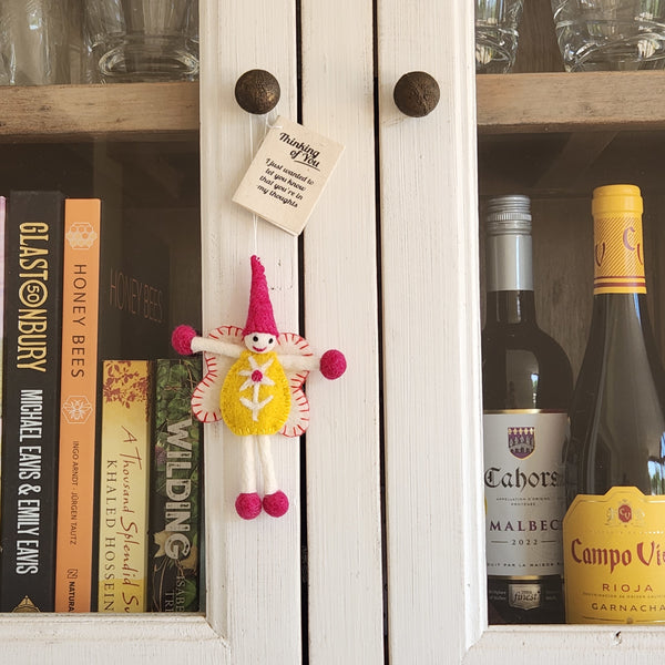 A small handmade felt fairy/pixie with a pink hat, yellow dress, and pink pom-pom limbs hangs on a white cabinet door.