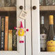 A small handmade felt fairy/pixie with a pink hat, yellow dress, and pink pom-pom limbs hangs on a white cabinet door.