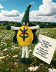 A handmade felt flower fairy keepsake stands in a grassy field with daisies under a blue sky. The ornament has a yellow felt flower body with green and purple details, a green pointed hat, and white felt limbs.