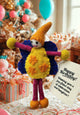 A handmade felt birthday doll with a yellow body, orange pointed hat, blue wings, and purple legs, holding a 'Happy Birthday' card in a decorated party setting with wrapped gifts and balloons.