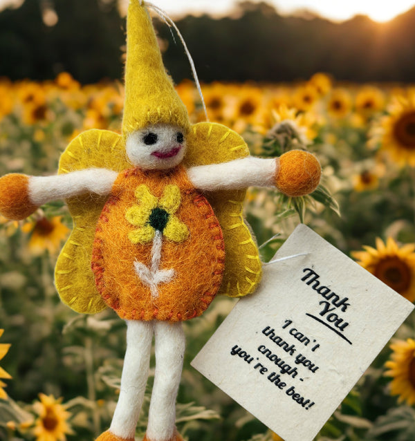 A handmade felt fairy sunflower doll ornament with a yellow hat and wings, orange flower body, and white stem/legs, photographed outdoors in a sunflower field. A small thank-you card is attached near the bottom right.