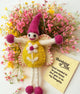 A handmade felt flower fairy doll with a pink pointed hat, yellow felted body, white wings with magenta stitching, and pink felted hands and feet. The doll is styled on a bed of small pink and yellow wildflowers next to a paper card that reads 'Thinking of You'.