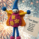 A handmade felt ornament shaped like a celebrating elf or fairy, featuring a blue hat and legs, yellow arms, a pink dress with yellow flowers, and orange wings. A paper tag reads 'Well Done! You did it! Woo hoo! Hooray! Congrats! High five!'.