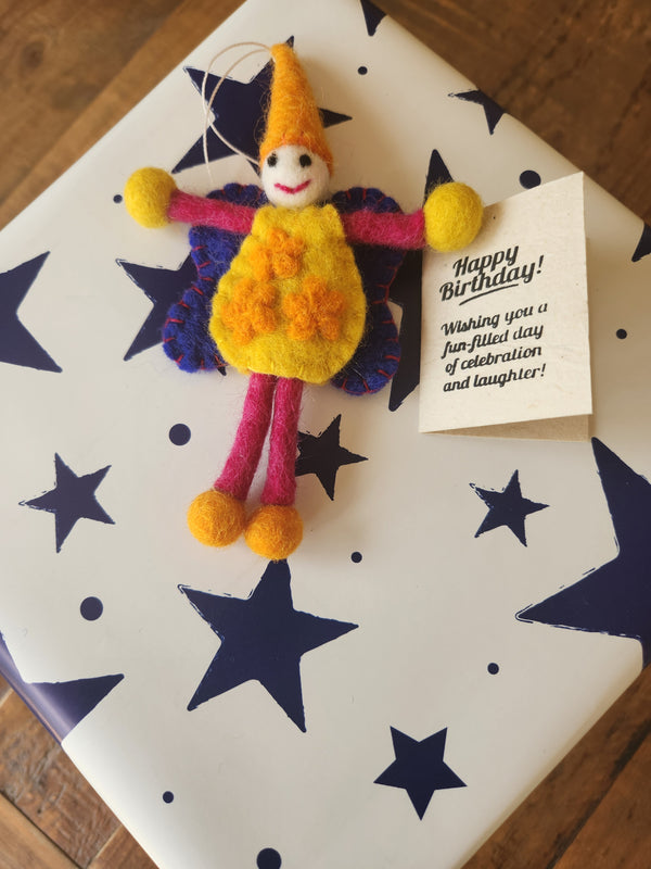 A handmade felt birthday message tag featuring a colorful fairy with yellow, pink, and purple details, placed on top of a white gift box decorated with navy blue stars. A small gift tag next to the doll reads 'Happy Birthday!'.