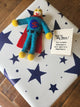 A handmade felt Superhero gift tag with a red cape and blue suit lies on top of a white gift box decorated with navy blue stars. A small card next to it reads 'You're My Hero! You're more than just a hero, you're a Superhero!'.