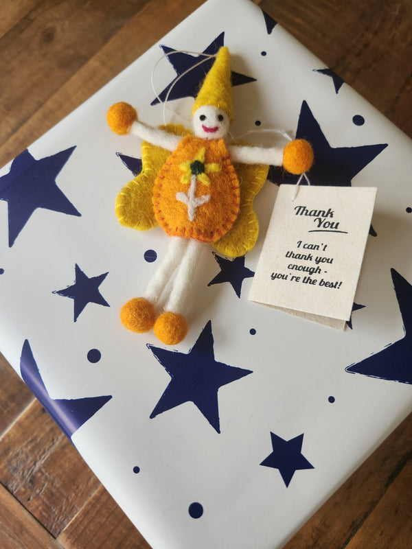 A handmade felt fairy/pixie gift tag with a yellow hat, orange body, white legs, and orange pom-poms, placed on top of a white gift box with dark blue stars. A small tag reads, 'Thank You I can't thank you enough, you're the best!'.