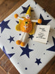 A handmade felt fairy/pixie gift tag with a yellow hat, orange body, white legs, and orange pom-poms, placed on top of a white gift box with dark blue stars. A small tag reads, 'Thank You I can't thank you enough, you're the best!'.
