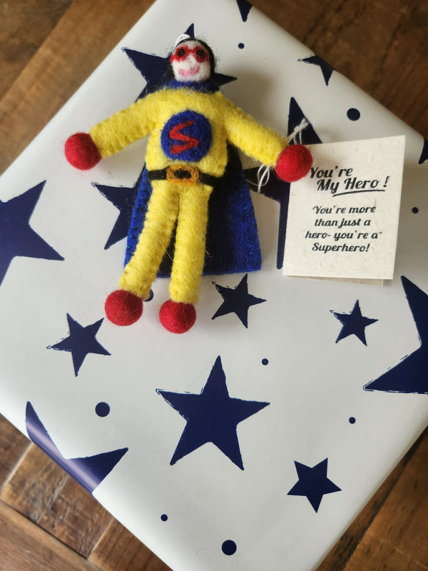 A handmade felt superhero gift tag/keepsake with a white face, red hands and feet, and a blue cape, placed on top of a white gift box with blue stars. A small card next to it reads, 'You're My Hero! You're more than just a hero - you're a superhero!'.