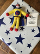 A handmade felt superhero gift tag/keepsake with a white face, red hands and feet, and a blue cape, placed on top of a white gift box with blue stars. A small card next to it reads, 'You're My Hero! You're more than just a hero - you're a superhero!'.