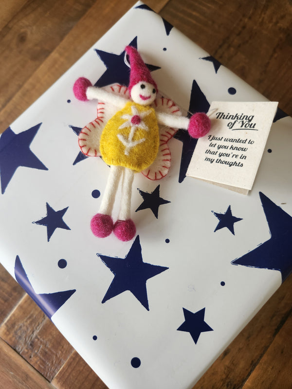 A handmade felt gift tag featuring a small whimsical fairy keepsake with pink and yellow colours, placed on top of a white gift box decorated with navy blue stars. A small tag next to it reads 'Thinking of You'.
