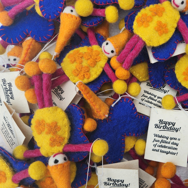 A close-up pile of handmade felt birthday keepsakes in bright blue, yellow, orange, and pink, each with a small white face and a white paper tag printed with a Happy Birthday message.