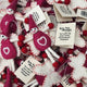 A pile of handmade felt Valentine's Day keepsake gift tags. The ornaments are made from pink and white felt, featuring small smiling faces, pointed hats, and a white heart outline on the front.