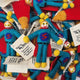 A pile of handmade felt super hero gift tags/keepsakes, featuring bright blue bodies, red capes, yellow pom-pom shoes and hands, and white paper tags with "You're My Hero" gift message.