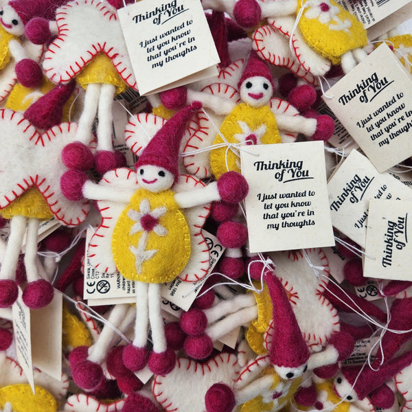 A pile of handmade felt fairy/pixie keepsakes/gift tags, each with a white tag that reads 'Thinking of You'. The ornaments feature yellow felt dresses, pink felt hats and pom-poms, and white felt bodies and legs.