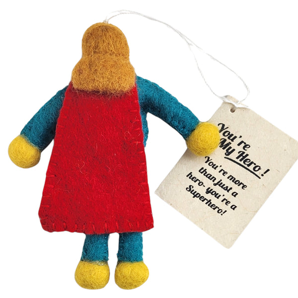 Th back of a handmade felt super hero figure keepsake ornament with a red cape, blue sleeves and legs, yellow hands and feet, and brown/blonde long felt hair. It is shown with an attached gift tag that reads 'You're My Hero! You're more than just a hero- you're a Superhero!'.
