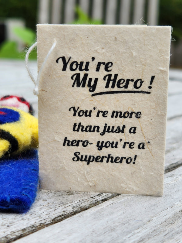 A handmade paper tag with black printed text reading 'You're My Hero! You're more than just a hero- you're a Superhero!' resting on a weathered white wooden table.