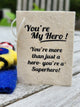 A handmade paper tag with black printed text reading 'You're My Hero! You're more than just a hero- you're a Superhero!' resting on a weathered white wooden table.