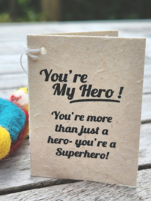 A handmade paper greeting card with black printed text reading 'You're My Hero! You're more than just a hero- you're a Superhero!' resting on a weathered wooden surface.