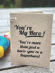 A handmade paper greeting card with black printed text reading 'You're My Hero! You're more than just a hero- you're a Superhero!' resting on a weathered wooden surface.