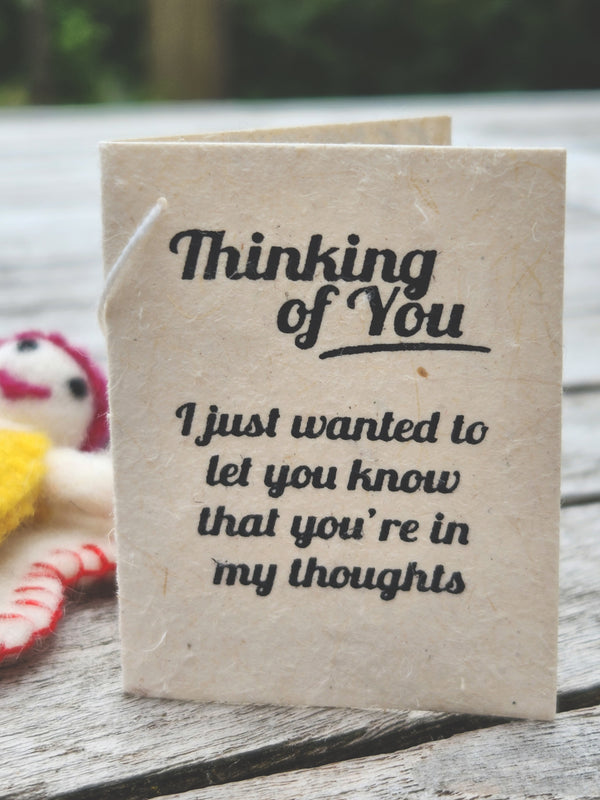A handmade paper greeting card/tag with black printed text reading 'Thinking of You. I just wanted to let you know that you're in my thoughts', standing on a weathered wooden surface.