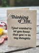 A handmade paper greeting card/tag with black printed text reading 'Thinking of You. I just wanted to let you know that you're in my thoughts', standing on a weathered wooden surface.
